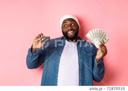 Dreamy african-american man showing credit card and dollars, thinking about shopping and smiling, standing over pink background 72870510