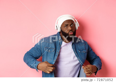 Cool and sassy Black guy dancing, listening to music in headphones, looking confident, standing over pink background 72871052