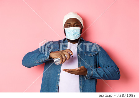 Covid-19, lifestyle and lockdown concept. Smiling african-american man in face mask cleaning hands with sanitizer, using antiseptic and looking at camera, pink background Covid-19, lifestyle and lockdown concept. Smiling african-american man in face mask cleaning hands with sanitizer, using antiseptic and looking at camera, pink background 72871083
