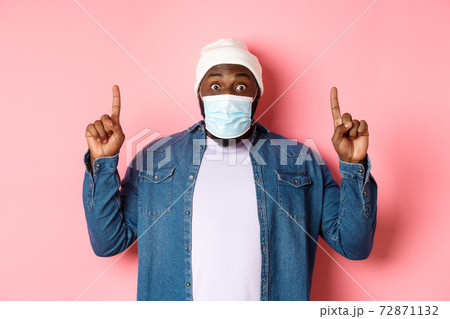 Coronavirus, lifestyle and global pandemic concept. Excited african-american guy in medical mask showing announcement, pointing fingers up, standing over pink background Coronavirus, lifestyle and global pandemic concept. Excited african-american guy in medical mask showing announcement, pointing fingers up, standing over pink background 72871132