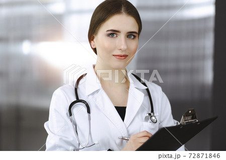 Friendly female doctor standing and holding clipboard in modern clinic. Portrait of cheerful smiling physician Friendly female doctor standing and holding clipboard in modern clinic. Portrait of cheerful smiling physician 72871846
