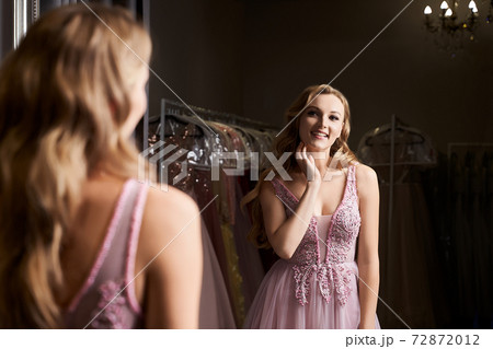 Young beautiful blonde girl wearing a full-length pink violet or purple chiffon prom ball gown decorated with sparkles and sequins. Model in front of mirror in a fitting room at dress hire service. Young beautiful blonde girl wearing a full-length pink violet or purple chiffon prom ball gown decorated with sparkles and sequins. Model in front of mirror in a fitting room at dress hire service. 72872012