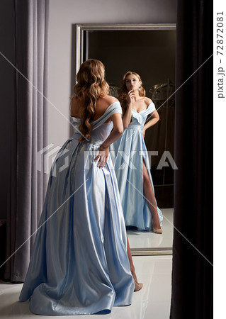 Young beautiful blonde girl wearing an off-the-shoulder full-length sky blue satin slit prom ball gown. Model looking in mirror. Fitting room in dress hire service. Young beautiful blonde girl wearing an off-the-shoulder full-length sky blue satin slit prom ball gown. Model looking in mirror. Fitting room in dress hire service. 72872081