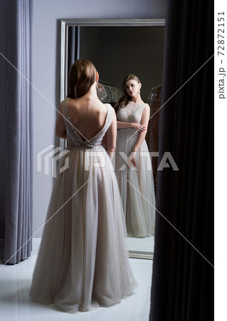 Young beautiful brunette girl wearing a full-length silver white chiffon prom ball gown decorated with sparkles and sequins. Model in front of mirror in a fitting room at dress hire service. 72872151