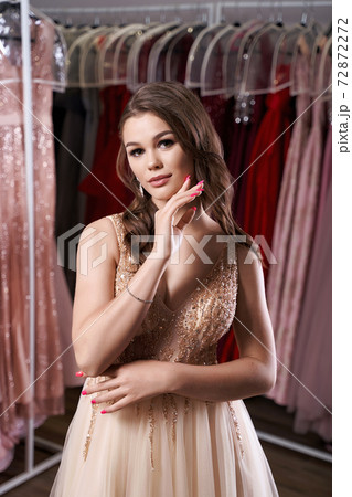 Young beautiful brunette girl wearing a full-length beige yellow champaign chiffon slit prom ball gown decorated with golden sparkles and sequins. Dress hire service with many dresses on background. 72872272
