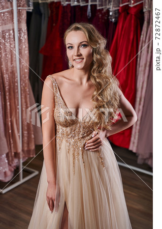 Beautiful blonde girl wearing a full-length yellow champaign chiffon slit prom ball gown decorated with golden sequins. Model in dress hire service with many dresses in different color and style. 72872467