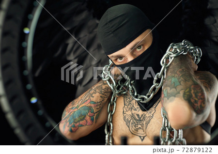portrait of a guy in a balaclava with a bare torso in tattoos holding chains in his hands 72879122