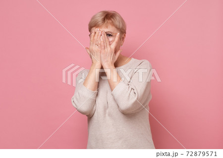 Pretty mature woman covering face with hands and peeping through her fingers at camera Pretty mature woman covering face with hands and peeping through her fingers at camera 72879571