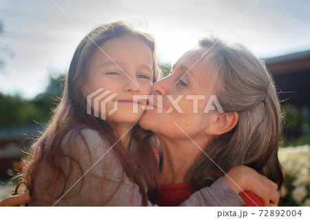 Senior grandmother with gray hair wearing red sweater with her little granddaughter are hugging in the garden and during sunny day outdoors, mother's day 72892004