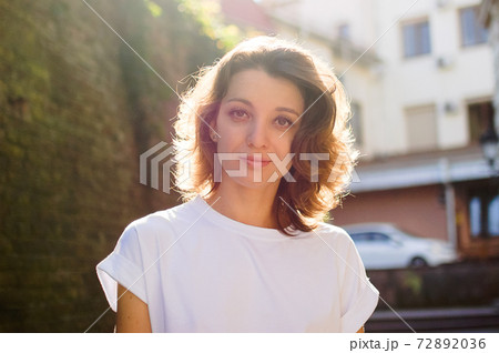 Female portrait of beautiful student, pretty girl with short hair in white t-shirt spending time outdoors near old stone wall during sunny spring or summer day 72892036