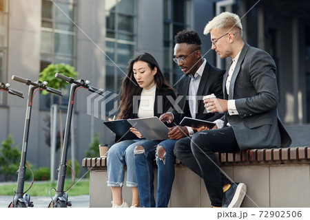 Elegant multiracial colleagues sitting on the bench near office and discussing business matters outdoors. Elegant multiracial colleagues sitting on the bench near office and discussing business matters outdoors. 72902506