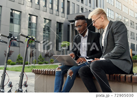 Elegant multiracial colleagues sitting on the bench near office and discussing business matters outdoors. Elegant multiracial colleagues sitting on the bench near office and discussing business matters outdoors. 72905030