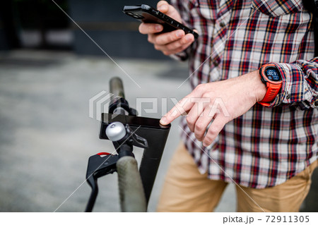 Male hands modern electric scooter turns on and tunes. Caucasian man dressed stylishly casual uses cell phone to control kick scooter, close-up no face. Personal electric transport in city 72911305