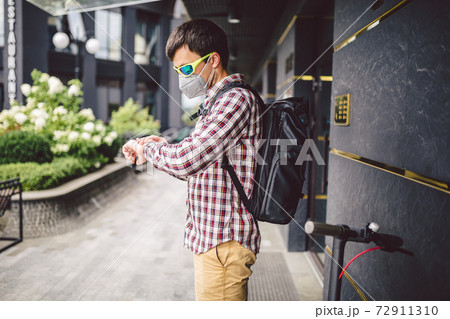 Courier uses electric scooter in city. Young attractive Caucasian man with backpack, stylishly dressed, using mask against smog and air pollution in city, waiting customer and looking at wristwatch 72911310
