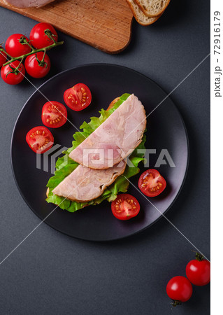 Sandwich with turkey ham meat, green salad and fresh cherry tomatoes slices on black plate near to ingredients on cutting board, grey minimal background, top view Sandwich with turkey ham meat, green salad and fresh cherry tomatoes slices on black plate near to ingredients on cutting board, grey minimal background, top view 72916179
