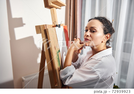 Young beautiful female artist wearing white shirt stands in front of easel and looks down 72939923