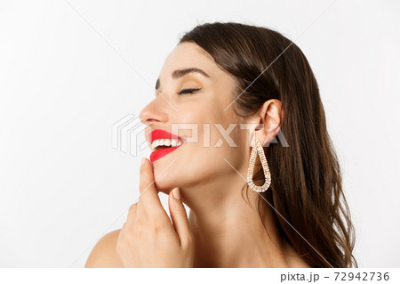 Fashion and beauty concept. Headshot of sensual brunette woman with earrings and red lipstick, smiling tempted and touching lip, standing over white background Fashion and beauty concept. Headshot of sensual brunette woman with earrings and red lipstick, smiling tempted and touching lip, standing over white background 72942736