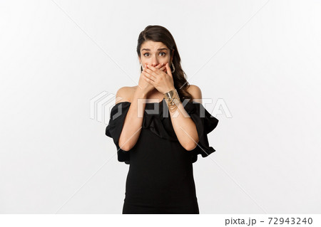 Fashion and beauty. Attractive woman in black dress cover mouth and gasping shocked, staring at camera worried, standing over white background Fashion and beauty. Attractive woman in black dress cover mouth and gasping shocked, staring at camera worried, standing over white background 72943240