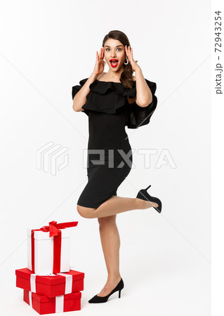 Surprised and happy woman standing in luxury dress and red lips, receive holiday presents and looking amazed, rejoicing from gifts, standing over white background Surprised and happy woman standing in luxury dress and red lips, receive holiday presents and looking amazed, rejoicing from gifts, standing over white background 72943254