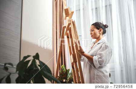 Beautiful woman painter with gathered hair in a bun and paint brushes in her hair standing in front of the easel in workshop Beautiful woman painter with gathered hair in a bun and paint brushes in her hair standing in front of the easel in workshop 72956458