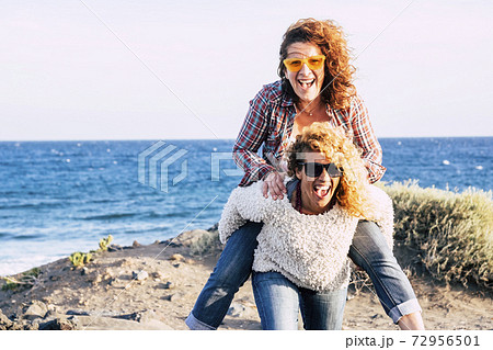 Happy and cheerful middle age couple of caucasian women enjoy and play together carrying eachother and laughing a lot - people having fun in outdoor leisure activity - concept of vacation with friends 72956501