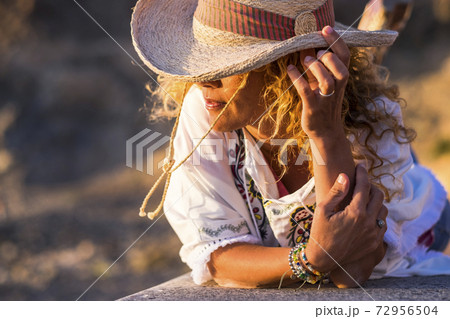 Portait of beautiful young trendy woman with cowboy hat and accessories like bracelets lay down on a wall bench enjoying the sunset light - people in outdoor leisure activity and vacation time 72956504