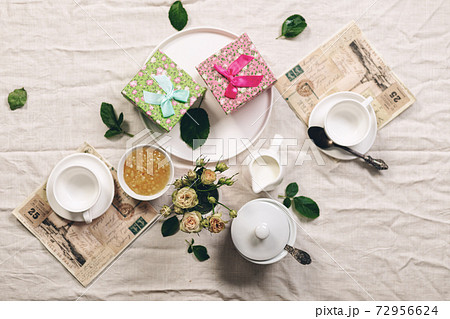 Food still life in french style. White coffee cups, milk jug with cream, roses and two gifts on a linen tablecloth. Refined toning. Top view Food still life in french style. White coffee cups, milk jug with cream, roses and two gifts on a linen tablecloth. Refined toning. Top view 72956624