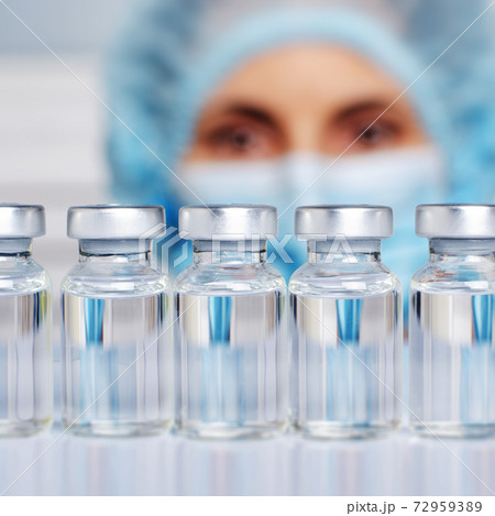 Female nurse in safety mask and cap looks at vials with liquid medicine on refrigerator shelf 72959389