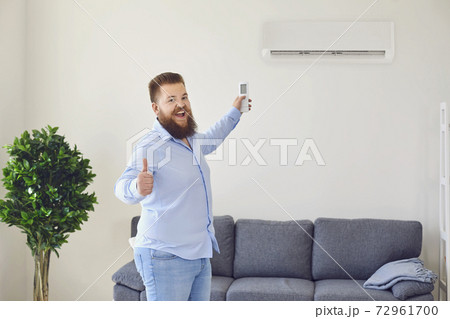 Funny bearded man turning on air conditioner with remote control and showing thumb up in apartment, copy space 72961700