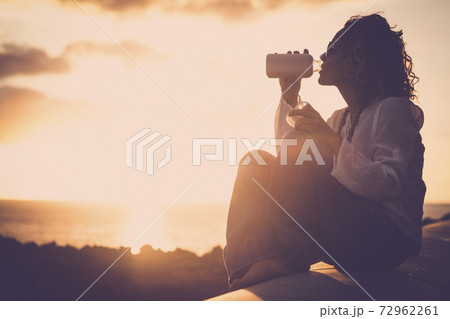 Happy and free woman alone enjoying the sunset on the coast with ocean view drinking a fresh drink - golden hour and vacation summer concept lifestyle for people 72962261