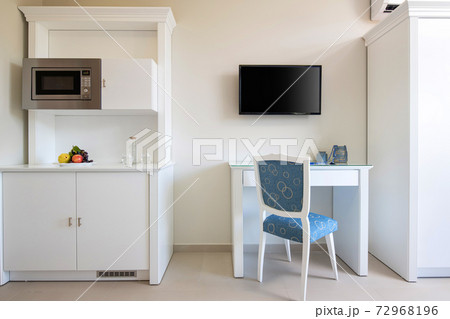 Front view of white wall with TV, table, classic blue chair, wooden kitchen cupboard cabinet with built-in appliances in small room modern interior Front view of white wall with TV, table, classic blue chair, wooden kitchen cupboard cabinet with built-in appliances in small room modern interior 72968196