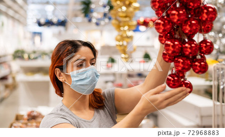 A woman in a mask chooses balls of decorations for a Christmas tree in the store A woman in a mask chooses balls of decorations for a Christmas tree in the store 72968883