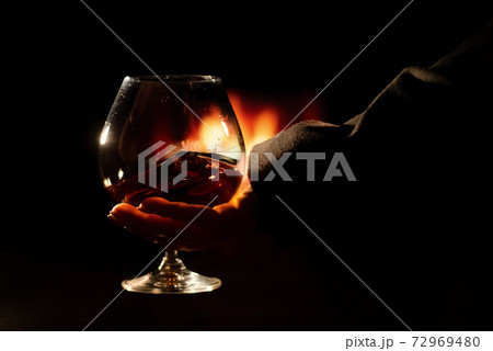 Close-up of a female hand with a glass of brandy on the background of the fireplace Close-up of a female hand with a glass of brandy on the background of the fireplace 72969480