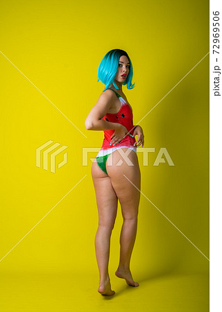 Beautiful woman in a swimsuit with a picture of a watermelon is standing with her back. Stylish girl in a colored short wig posing in the studio on a yellow background. 72969506