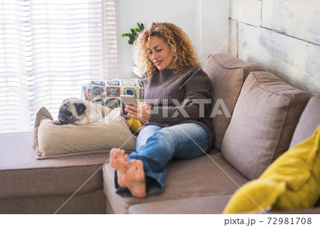 Happy adult beautiful curly woman at home typing and reading from celllar phone with her best friend dog pug relaxing at her side - concept of lfiestyle for relaxed hapy independent people Happy adult beautiful curly woman at home typing and reading from celllar phone with her best friend dog pug relaxing at her side - concept of lfiestyle for relaxed hapy independent people 72981708
