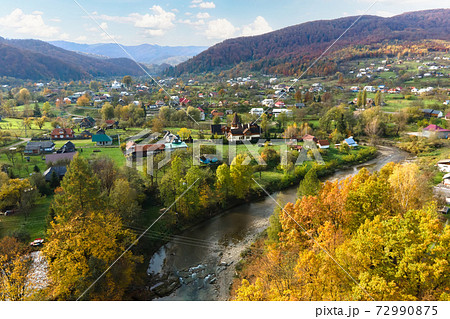 Aerial view of a village rural area with small houses between autumn mountain hills covered with yellow and green spruce forest. 72990875