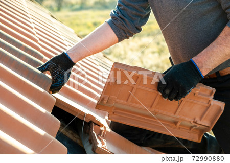 Closeup of worker hands installing yellow ceramic roofing tiles mounted on wooden boards covering residential building roof under construction. 72990880