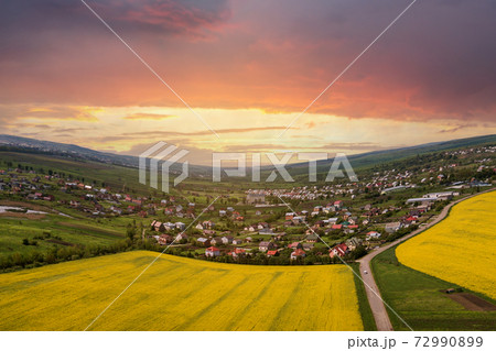 Aerial view of ground road with moving cars in green fields with blooming rapeseed plants, suburb houses on horizon and blue sky copy space background. Drone photography. Aerial view of ground road with moving cars in green fields with blooming rapeseed plants, suburb houses on horizon and blue sky copy space background. Drone photography. 72990899