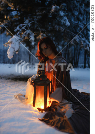 Young woman sitting under fir tree covered in snow looking at vintage lantern. Medieval fairytale concept Young woman sitting under fir tree covered in snow looking at vintage lantern. Medieval fairytale concept 73010805