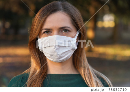Young woman wearing white cotton virus mouth nose mask, nice bokeh - blurred park trees - in background, closeup face portrait Young woman wearing white cotton virus mouth nose mask, nice bokeh - blurred park trees - in background, closeup face portrait 73032710