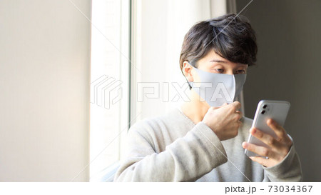 窓際でスマホを操作するマスク装着の若い男性 窓際でスマホを操作するマスク装着の若い男性 73034367