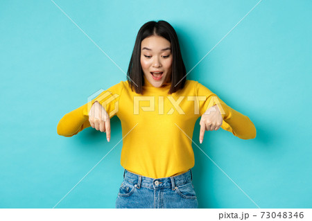 Shopping concept. Surprised cute asian girl checking out discounts, pointing fingers down and looking amazed, saying wow, standing over blue background 73048346