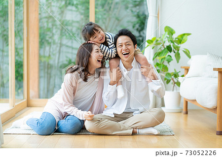 リビングに座ってパパに肩車をされてふざける女の子とパパとママの3人家族のポートレート 73052226