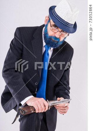 An elderly man in glasses with a blue beard holds a huge knife in his hands and looks frighteningly from under his foreheads 73052864