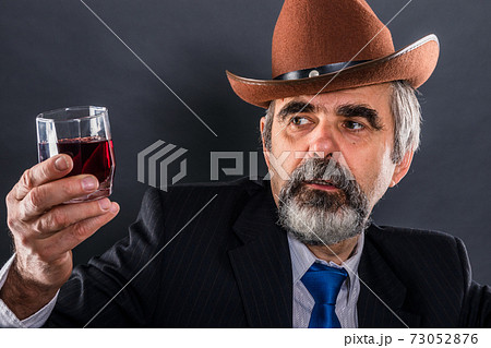 Elderly man in suit with tie and cowboy hat evaluates the transparency of wine 73052876