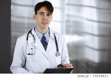 Friendly young male doctor standing with clipboard in modern clinic. Medicine concept Friendly young male doctor standing with clipboard in modern clinic. Medicine concept 73059621
