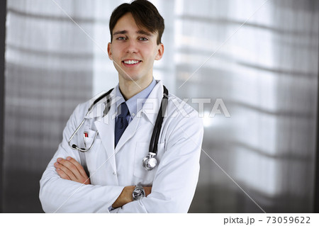 Cheerful smiling man-doctor standing with arms crossed in clinic. Perfect medical service with young smart physician in hospital. Medicine concept Cheerful smiling man-doctor standing with arms crossed in clinic. Perfect medical service with young smart physician in hospital. Medicine concept 73059622