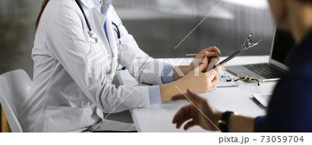 Unknown young woman doctor is listening to her patient, while sitting together in clinic. Medicine concept during Coronavirus pandemic 73059704
