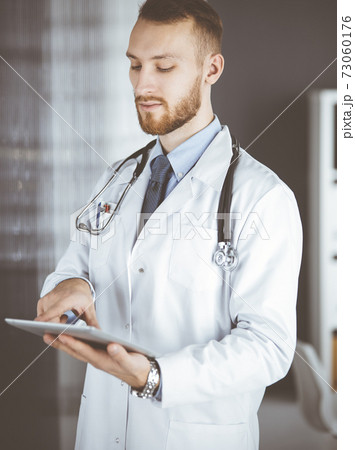Friendly red-bearded doctor using tablet computer in clinic at his working place. Perfect medical service in hospital and medicine concept 73060176