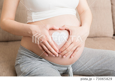a pregnant girl sits at home on the bed and smears heart an anti-stretch mark cream on her stomach. Pregnancy, motherhood, preparation and expectation concept 73065829